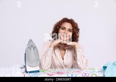 Eine lächelnde Frau sitzt am Bügelbrett und am Bügeleisen. Stockfoto