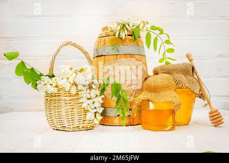 Frische flüssige Akazienhonigernte in einem Holzfass und einem transparenten Glas mit einer Leinenserviette auf dem Deckel. Zweige blühender weißer Akazie, Rotkehlchen Stockfoto