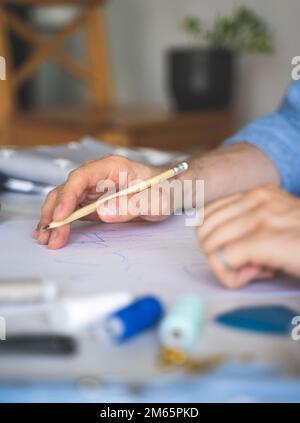 Die Hände des selbst unterrichteten Designers sind auf seiner Zeichnung. Hand mit Bleistift. DIY-Designer zeichnet eine Skizze für ein neues Kostümprojekt. Nahaufnahme, selektiver Fokus Stockfoto