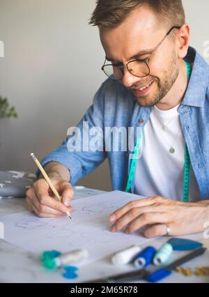 Seamster mit Brille arbeitet am Tisch.DIY-Designer zeichnet eine Skizze für ein neues Kostümprojekt.lächelnder Mann in einem blauen Hemd erstellt eine Skizze Stockfoto