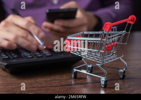 Online-Shopping-Konzept. Kleiner Supermarkt, Einkaufswagen zum Einkaufen, Ein Mann zählt die Kosten für Einkäufe mit einem Taschenrechner. Stockfoto