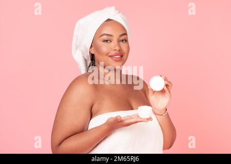 Hautpflegekonzept. Wunderschöne afroamerikanische Frau in Übergröße, die eine offene Cremedose hält und im pinkfarbenen Studiohintergrund posiert Stockfoto