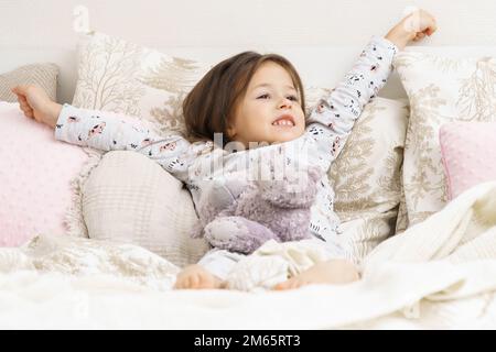 Glückliches kleines Mädchen in Schlafkleidung liegt auf dem Bett und streckt sich mit erhobenen Armen. Porträt eines erwachenden Kindes. Das Kind wacht morgens auf. Mit Füllung schlafen Stockfoto