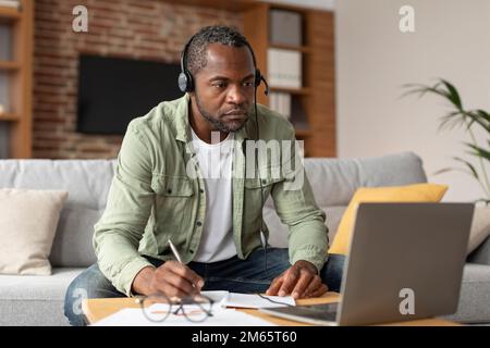 Neuer Normalbetrieb und neuer Job aus der Ferne. Ein fleißiger, reifer, schwarzer Mann mit Kopfhörern macht sich Notizen und sieht sich den Computer an Stockfoto