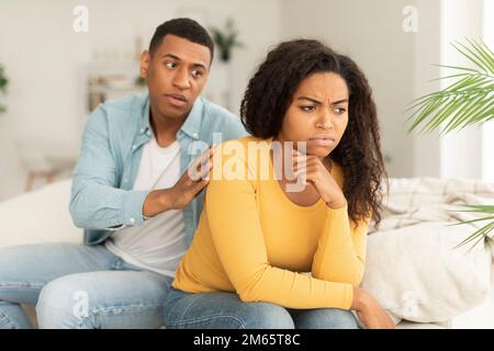 Trauriger Ehemann beruhigt verärgerte afroamerikanische Ehefrau, ignoriert Typen nach Streit im Wohnzimmer Stockfoto