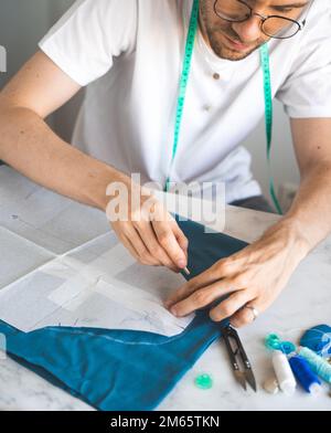 Ein selbstlernender Schneider in weißem T-Shirt und einer Brille arbeitet mit Stoff, Muster und Kreide. Männlicher Designer überträgt eine Vorlage in ein Stoffstudio Stockfoto