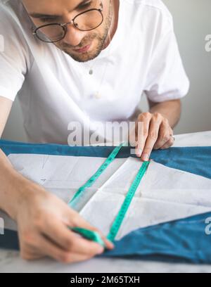 Ein Schneider verwendet ein Maßband während seiner Arbeit in der Werkstatt. Ein selbstlernender Näher in weißem T-Shirt und Brille funktioniert mit Stoff, Muster und ch Stockfoto