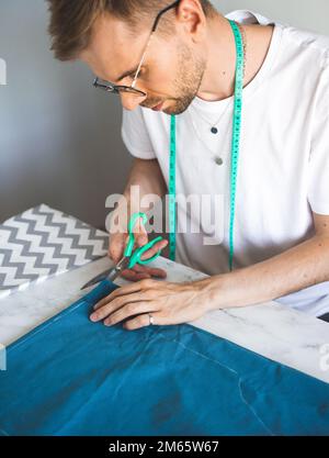 Ein selbstlernender Näher in weißem T-Shirt und einer Brille arbeitet mit Stoff, Muster und Kreide. Ein Schneider schneidet Stoff in einem Heimstudio. Kleine Unternehmen Stockfoto