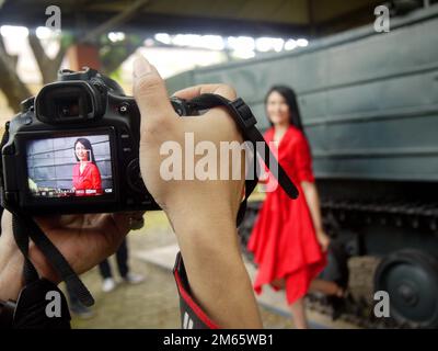 Ein Fotograf fotografiert ein indonesisches weibliches Modell. Das weibliche Model trägt ein rotes Kleid. Fotografen nehmen Bilder mit einer DSLR-Kamera auf. Stockfoto