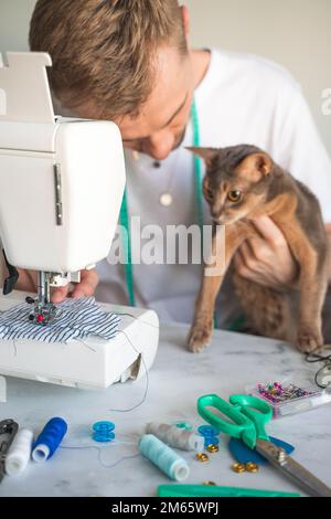 Nähkleidung für Haustiere. Katzenkleidung für Katzen. Ein lächelnder Näher näht Kleider für seine abyssinische Katze. Das Konzept eines kleinen Unternehmens Stockfoto