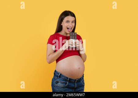 Porträt Einer Fröhlichen Jungen Schwangeren Frau, Die Schokolade Beißt, Mit Gelbem Hintergrund Stockfoto