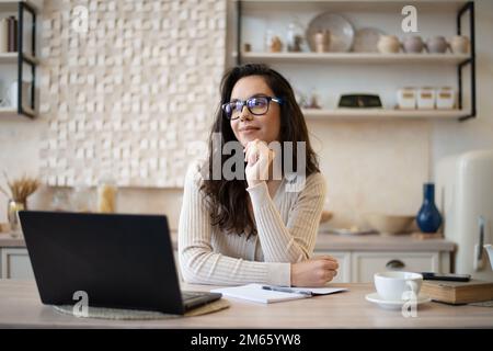 Eine traumhafte Frau sitzt in der Küche am Tisch mit einem geöffneten Laptop, blickt weg und denkt nach, Platz zum Kopieren Stockfoto