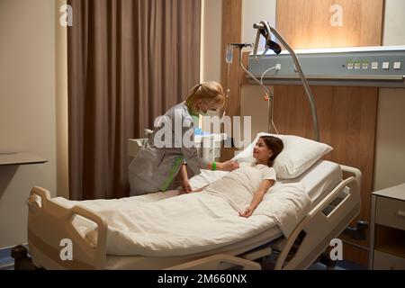 Eine Frau mit Schutzmaske lehnte sich auf ein Mädchen in einem Krankenhausbett Stockfoto