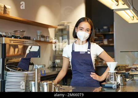 Covid-19 und Café-Arbeit. Asiatische Barista in Uniform, steht hinter der Theke, arbeitet als Barkeeper im Café, trägt medizinische Maske von Stockfoto