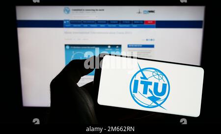 Person, die ein Mobiltelefon mit dem Logo der Internationalen Fernmeldeunion (ITU) auf dem Bildschirm vor der Webseite hält. Konzentrieren Sie sich auf das Display des Telefons. Stockfoto