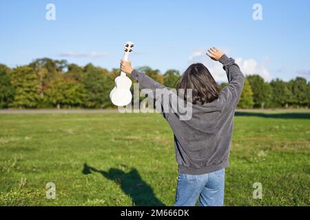 Fröhliche junge Frau, die mit ihrem Musikinstrument tanzt. Mädchen hebt ihre Ukulele hoch und posiert im Park auf grünem Feld Stockfoto