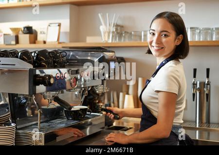 Ein lächelndes asiatisches Barista-Mädchen macht Cappuccino mit einer Kaffeemaschine, steht hinter der Theke im Café Stockfoto