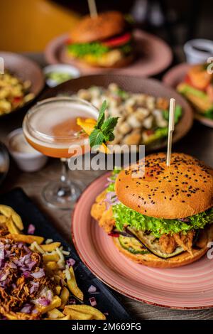 Vertikale von saftigen Grillhamburgern mit Spieß, Speisen und Getränken im Überfluss auf hölzernem Cafétisch. Pommes Frites, Salatplatte Stockfoto