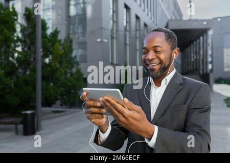 Ein afroamerikanischer Mann in einem Business-Anzug vor dem Bürogebäude, der sich ein Online-Video mit der App auf einem Tablet ansieht, ein Geschäftsmann lächelt und glücklich ist. Stockfoto