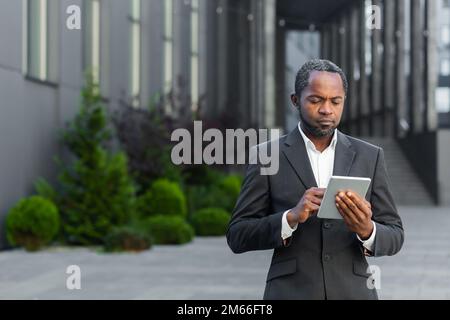 Ernsthaft denkender afroamerikanischer Geschäftsmann, der einen Tablet-Computer benutzt, Mann in einem Geschäftsanzug, der Nachrichten online liest und vor dem Bürogebäude steht. Stockfoto