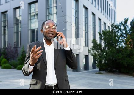 Fröhlicher und zufriedener afroamerikanischer Chef außerhalb des Bürogebäudes, lächelnd und telefonisch mit Kollegen, Mann im Geschäftsanzug, der in der Mittagspause spaziert. Stockfoto