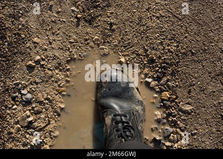 Ein dreckiger Militärstiefel voller Schlamm steht in einer Pfütze von Regen. Das abgenutzte Schuhwerk weist auf eine harte Nutzung und eine raue Umgebung hin. Schließen Stockfoto