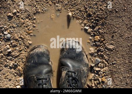 Zwei schmutzige Militärstiefel stehen in einer schlammigen Pfütze, was auf eine raue Umgebung und harte Nutzung hindeutet. Die Stiefel sind abgenutzt und mit Schlamm bedeckt. Schließen Stockfoto