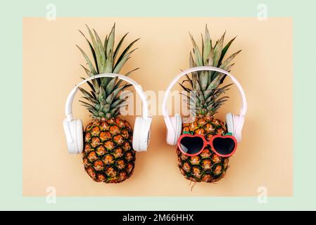 Zwei Ananas in Sonnenbrille mit Kopfhörern auf hellgrünem und gelbem Hintergrund. Sommerkonzept. Draufsicht, flach liegend. Stockfoto
