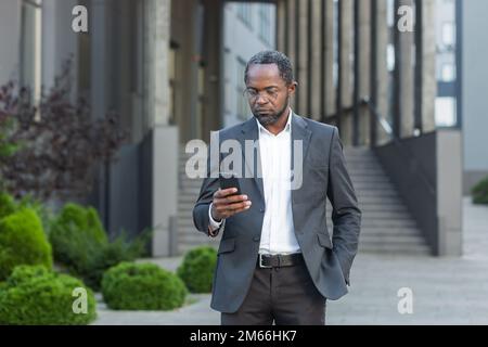 Seriöser und erfolgreicher afroamerikanischer Geschäftsmann außerhalb des Bürogebäudes mit Smartphone, Mann im Geschäftsanzug, der Nachrichten liest und online denkt und Internetseiten durchsucht. Stockfoto