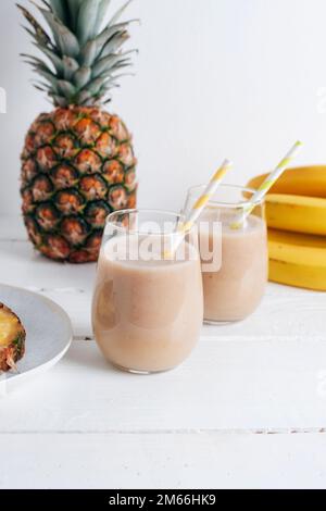 Zwei Gläser Bananen- und Ananas-Smoothie auf einem weißen Holztisch. Minimal-Entgiftungsdiät-Konzept, Sommer-Vitamingetränk. Nahaufnahme. Stockfoto