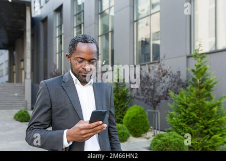 Seriöser und erfolgreicher afroamerikanischer Geschäftsmann außerhalb des Bürogebäudes mit Smartphone, Mann im Geschäftsanzug, der Nachrichten liest und online denkt und Internetseiten durchsucht. Stockfoto