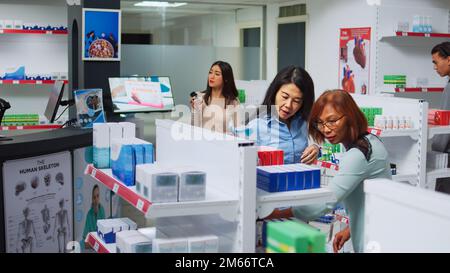 Eine Apothekerin, die asiatischen Klienten hilft, Medikamente zu finden, der Frau eine Pillen-Box gibt. Mitarbeiter schaut sich Supplemente und Medikamente in Regalen an und zeigt dem Kunden pharmazeutische Produkte. Stockfoto