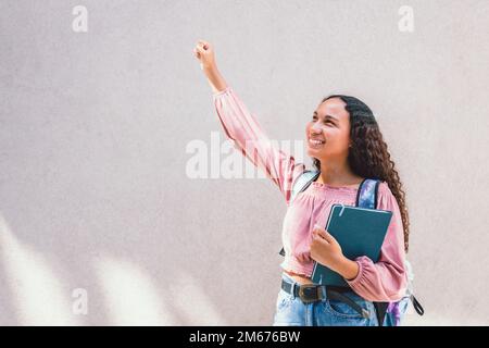 Lächelnde lateinische Studentin, die ihren Arm bei der Feier hebt. Vor einer Wand. Gesundes Selbstvertrauen Stockfoto