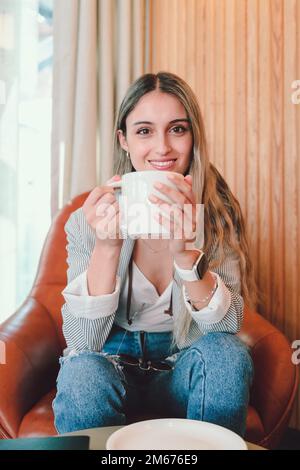 Lächelnde junge Frau, die auf einem Sessel sitzt, Kaffee aus einer Tasse trinkt und in die Kamera schaut Stockfoto