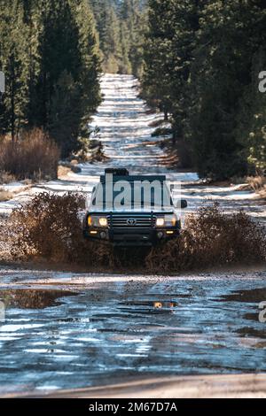 Vorderansicht des Off Road Overland Land Cruiser SUV, der das Wasser auf einem schneebedeckten Pfad im Wald durchquert Stockfoto