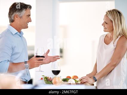Kochen und Konversation. Ein reifes Paar, das Wein genießt und sich unterhält, während es zusammen Abendessen macht. Stockfoto