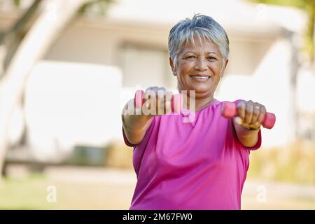 Hier wird man nicht alt. Porträt einer erwachsenen Frau, die draußen Kurzhanteln hebt. Stockfoto