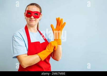 Junge Frau mit roter Maske und Schürze zieht Gummihandschuhe auf blauem Hintergrund an Stockfoto