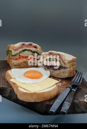 Hausgemachte Sandwichs mit gefülltem Schinken, Käse, Krabbenstäbchen und Gemüse mit Käse und gebratenem Ei auf Brot serviert mit Messer und Gabel auf Holzhintergrund. Stockfoto