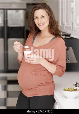 Ich hole ihr Vitamine aus Joghurt. Eine junge schwangere Frau, die in der Küche Joghurt isst. Stockfoto