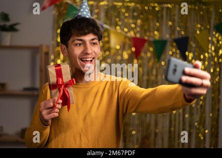 Junger glücklicher Mann mit Geschenkbox mit rotem Band, der zu Hause ein Mobiltelefon für Videoanrufe benutzt.Virtuelle Geburtstagsfeier mit Freunden oder Eltern.Online Stockfoto