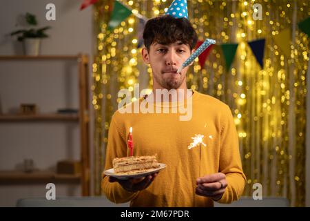 Ein fröhlicher junger Mann mit einem Springmütze und einem beleuchteten Funkel in den Händen feiert seinen Geburtstag, als er während der Feier im eine Kerze auf seinen Geburtstagskuchen blaste Stockfoto