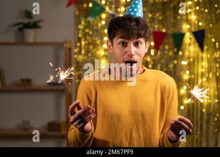 Ein gut aussehender Mann, der überraschend in die Kamera schaut und merkt, dass die Party vorbei ist, er ist spät dran. Ein schockierter Mann mit Geburtstagsmütze, der Sparkler im Arm hält Stockfoto