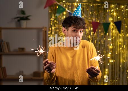 Ein junger Mann, der eine Geburtstagsparty-Mütze trug, war überrascht und schockiert, sah mit großer Zögerlichkeit in die Kamera, zuckte mit den Schultern, hielt die Glitzer drin Stockfoto