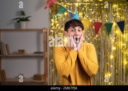 OH, MEIN GOTT! Nahaufnahme Foto eines arabers mit festlichem Hut, der gute Nachrichten hört, offene Mundarme auf den Wangen, lässiger gelber Pullover auf der Heim-Geburtstagsparty Stockfoto