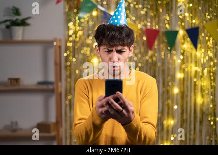 Porträt eines araber mit festlichem Hut, der Handys benutzt, um falsche schlechte Nachrichten zu lesen und seine Geburtstagsparty zu Hause zu feiern Stockfoto