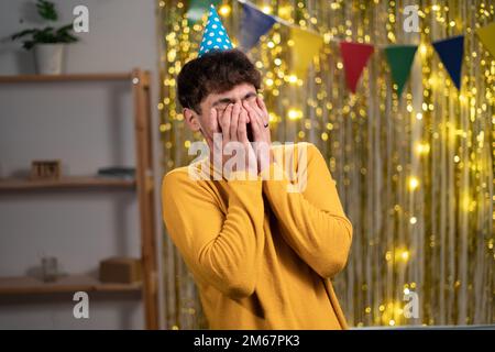 Aufgebracht, weinender junger Mann mit Partymütze, der seinen Geburtstag zu Hause feiert, unerwarteter Gast, Überraschung auf der Party Stockfoto