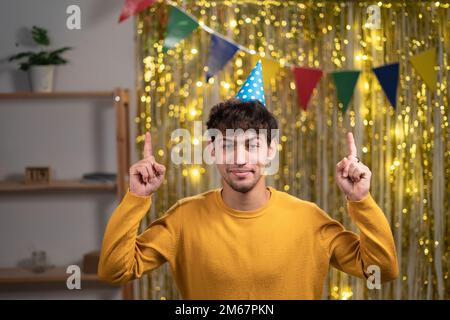 Ein Araber mit weihnachtlichem Hut, der Finger in den leeren Raum zeigt, Käufern niedrige Preise empfiehlt, und einen legeren gelben Pullover trägt, der seinen Geburtstag feiert Stockfoto