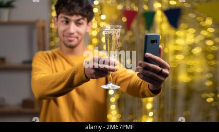 Ein junger Mann feiert seinen Geburtstag durch virtuelle Videoanrufe mit Freunden, die ein Smartphone verwenden. Glas mit Champagner halten. Authentisch dekoriert Stockfoto