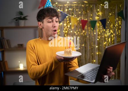 Virtuelle Geburtstagsfeier. Ein glücklicher Kerl mit Partyhut feiert online Geburtstag, bläst Kerze auf Kuchen, benutzt Laptop für Videogespräche mit Freunden oder Stockfoto